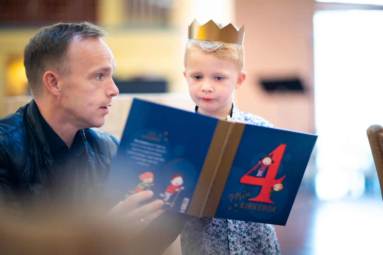 Bildet viser en fireåring med krone på hodet som ser i 4 års-boka sammen med pappan sin.