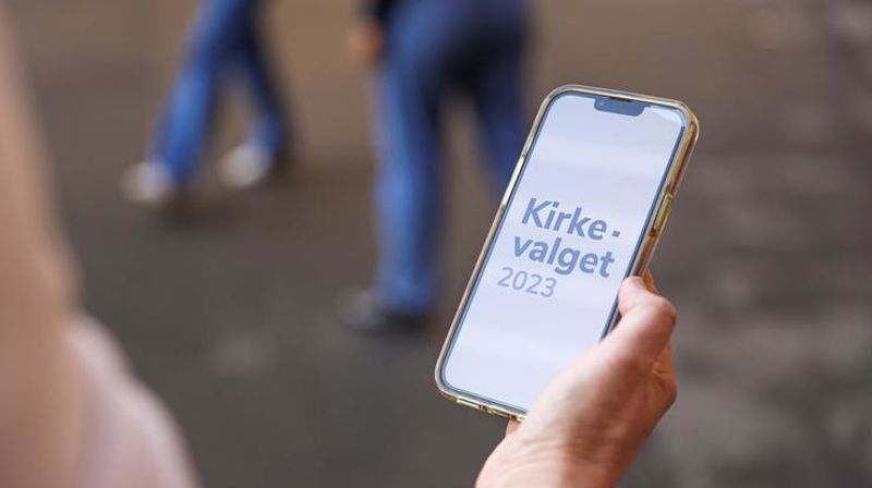 En hånd som holder en telefon hvor det står "Kirkevalget 2023"