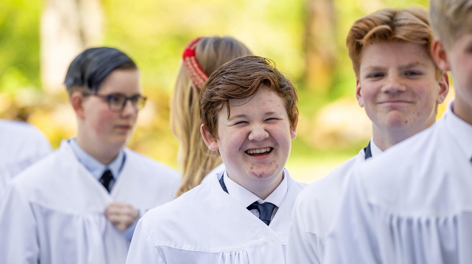 Mange ungdommer i hvite kapper klare for konfirmasjon, en av de smiler så bredt at øynene lukker seg