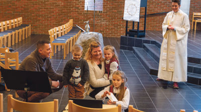 En familie som sitter foran en dataskjerm i kirkerommet. Det er dåp i kirken.