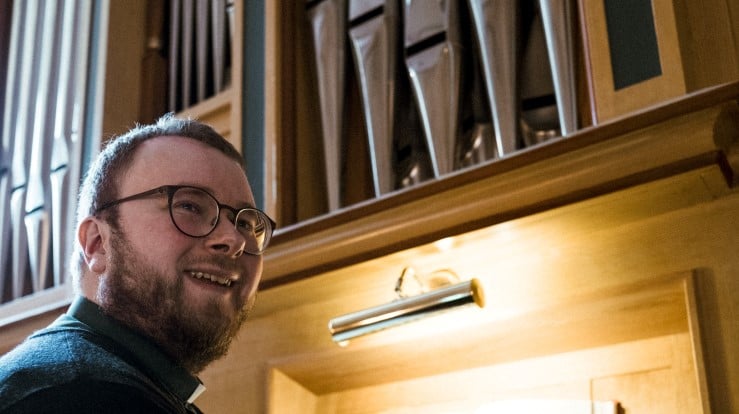 En kantor som sitter ved et orgel i et kirkerom. Han smiler.