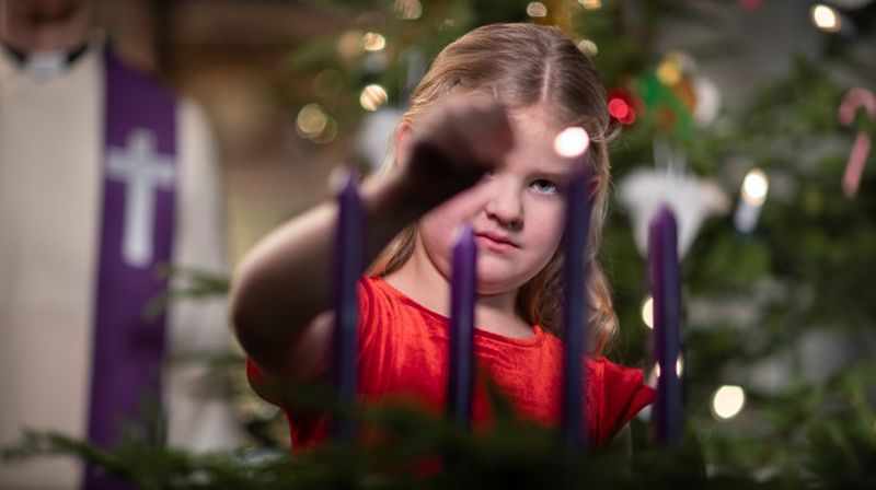 Ei jente tenner adventsstaken. Hun er pyntet i rød kjole. I bakgrunnen står et juletre. 