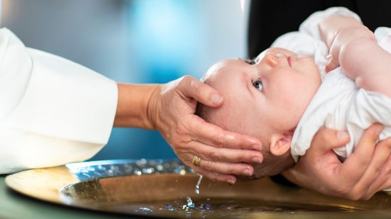 Det er dåp i kirken. Et barn blir holdt over en døpefont, og skal til å få vann over hodet.