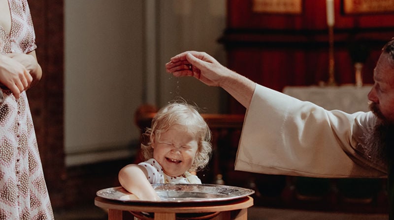 En treåring står ved døpefonten og leker med vannet. Presten sitter på huk ved siden av henne og døper henne med vann i pannen.