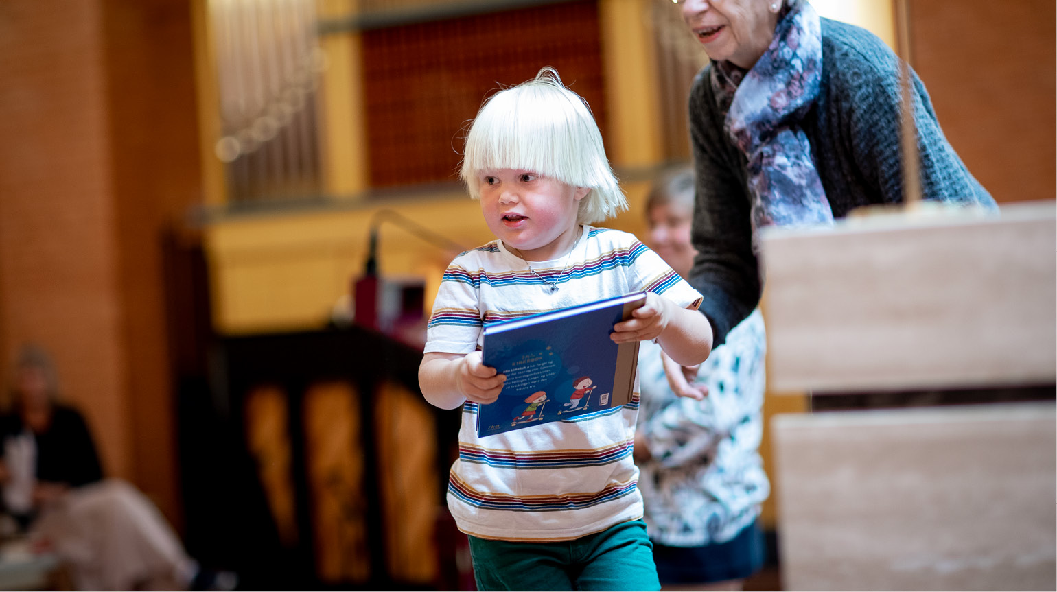 Gutt løper med 4årsbok i hendene. Han er inne i et kirkerom og det sitter andre rundt. Gutten smiler.