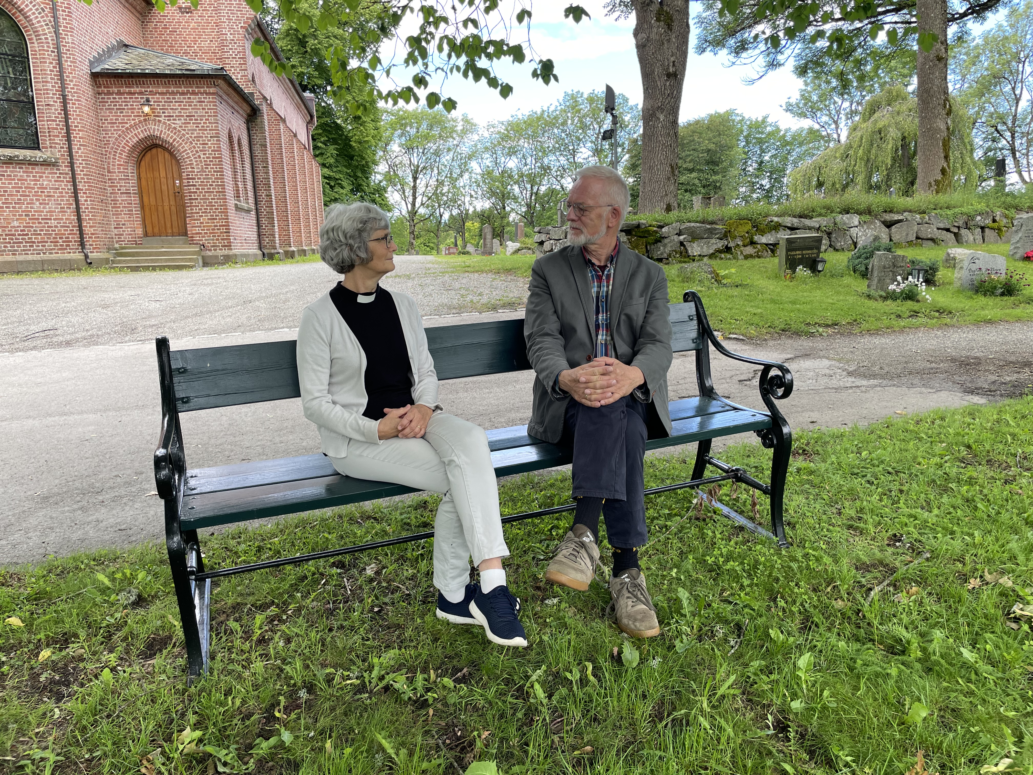 Synstolkning: prest og pensjonist i samtale, de sitter på en benk utenfor Ås kirke. Foto: Linda Janson-Haddal