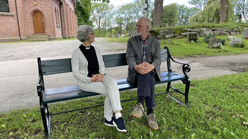 Synstolkning: prest og pensjonist i samtale, de sitter på en benk utenfor Ås kirke. Foto: Linda Janson-Haddal
