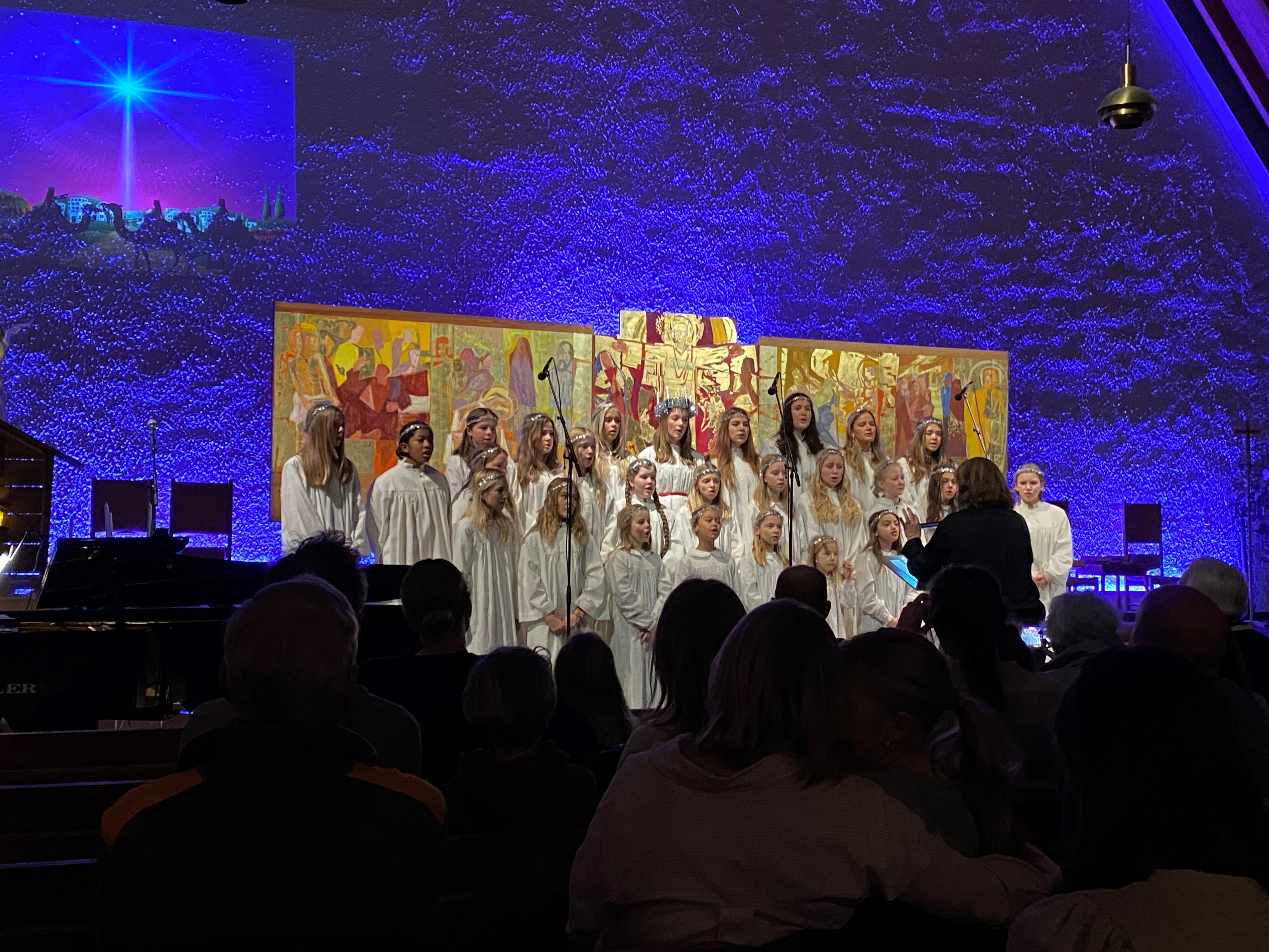 Korsangere i hvite kapper foran Holmen kirkes altertavle og blå fondvegg. 