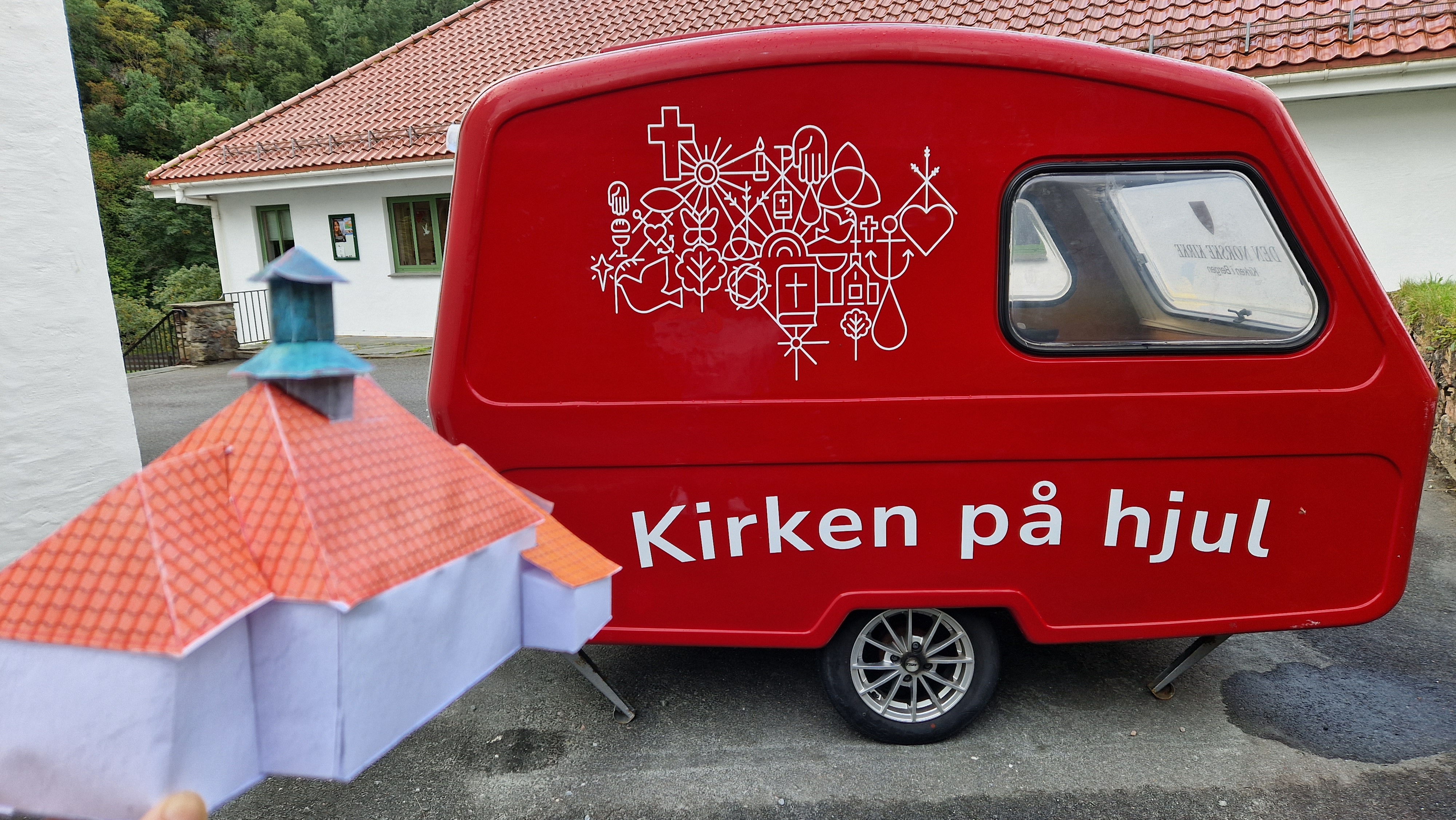 Rød, liten campingvogn og en pappmodell av en kirke. Foto utendørs.