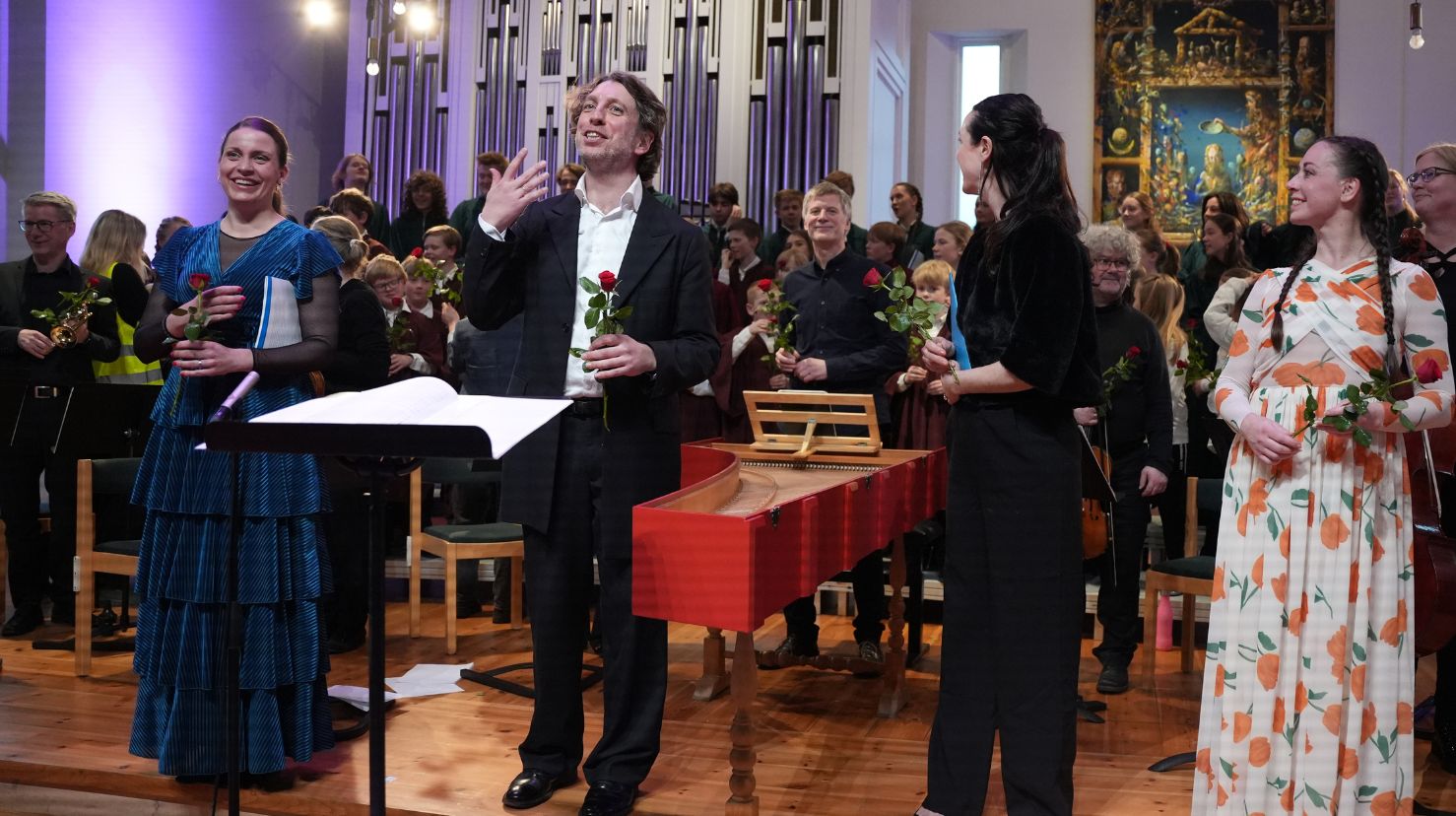 Fire pent kledde personer mottar applaus og rød rose foran et stort barnekor. Foto.