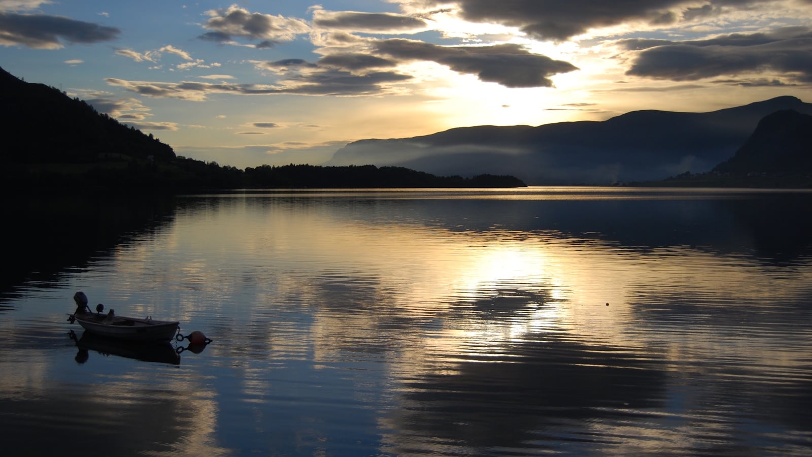 Solnedgang over fjorden ein jonsokkveld, ein liten båt ligg ute for anker, mørke fjell i bakgrunnen.