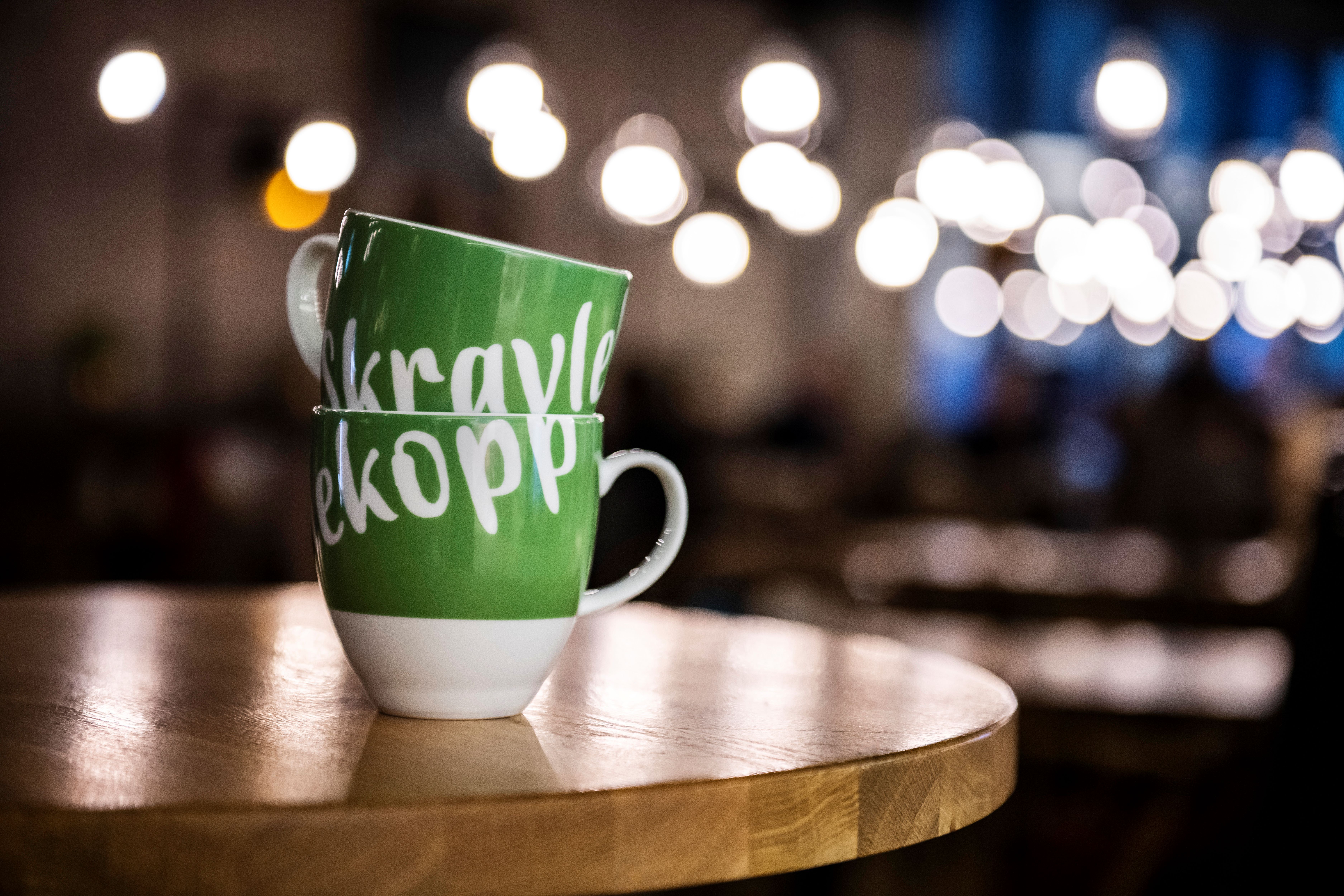 Bildet av en hvit og grønn kopp med teksten Skravlekopp på et cafebord.