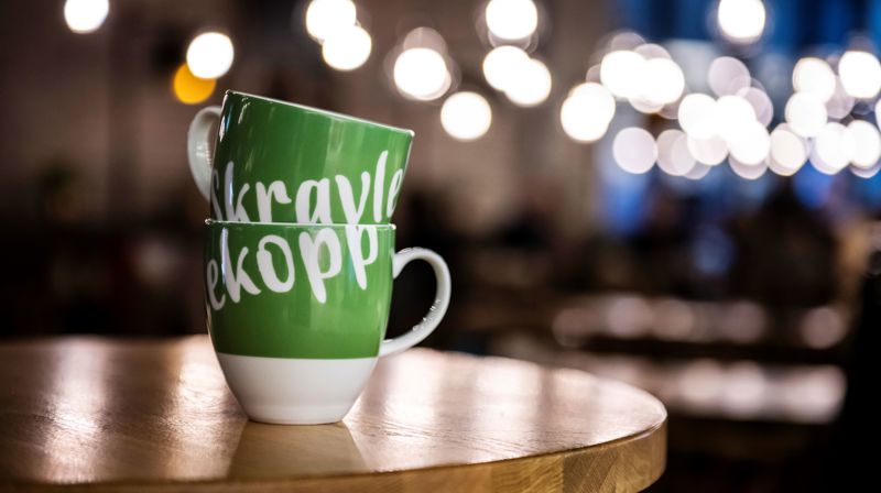 Bildet av en hvit og grønn kopp med teksten Skravlekopp på et cafebord.