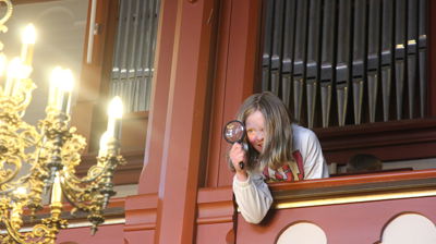 Bilde av en jente på orgelgalleriet i kirka. Hun kikker gjennom et forstørrelsesglass.