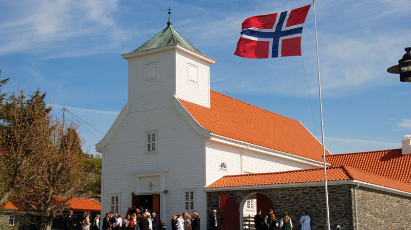 Blomvåg kyrkje mot blå himmel. Flagge er heist og strekk seg i vinden. Festkledde mennesker står utanfor kyrkja.