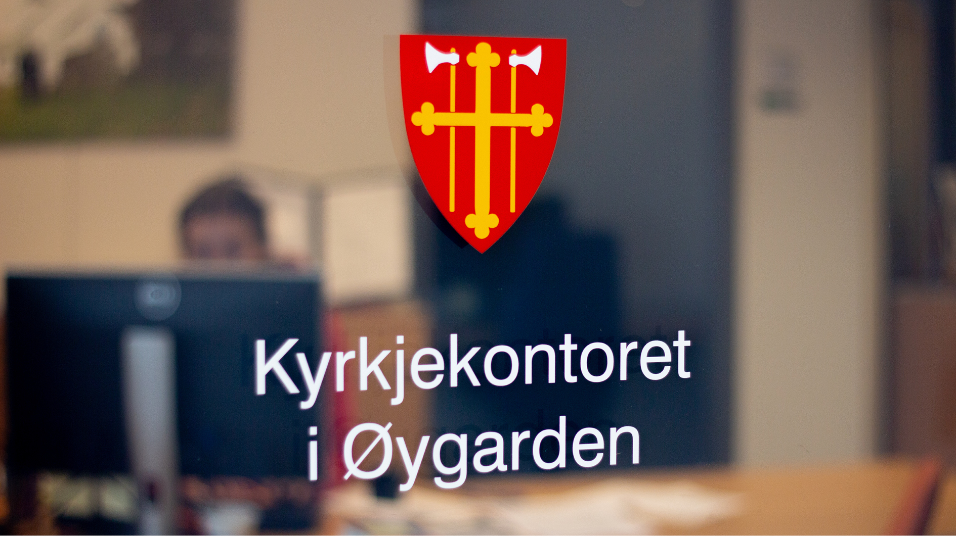Logoen til Den norske kyrkja, rødt våpenskjold med gult kors og økser, på døren inn til kyrkjekontoret. Man kan skimte at det sitter noen i resepsjonen i bakggrunnen. 