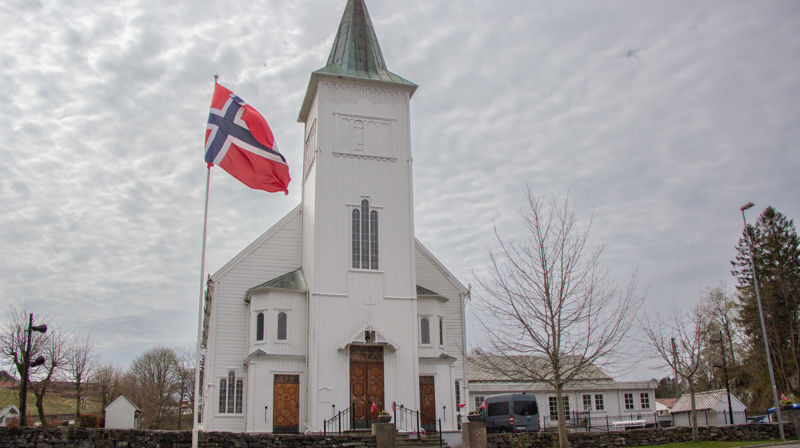 Fjell kyrkje i grå-ver, flagget er heist og det er pynta med småflagg i blomsterkassane