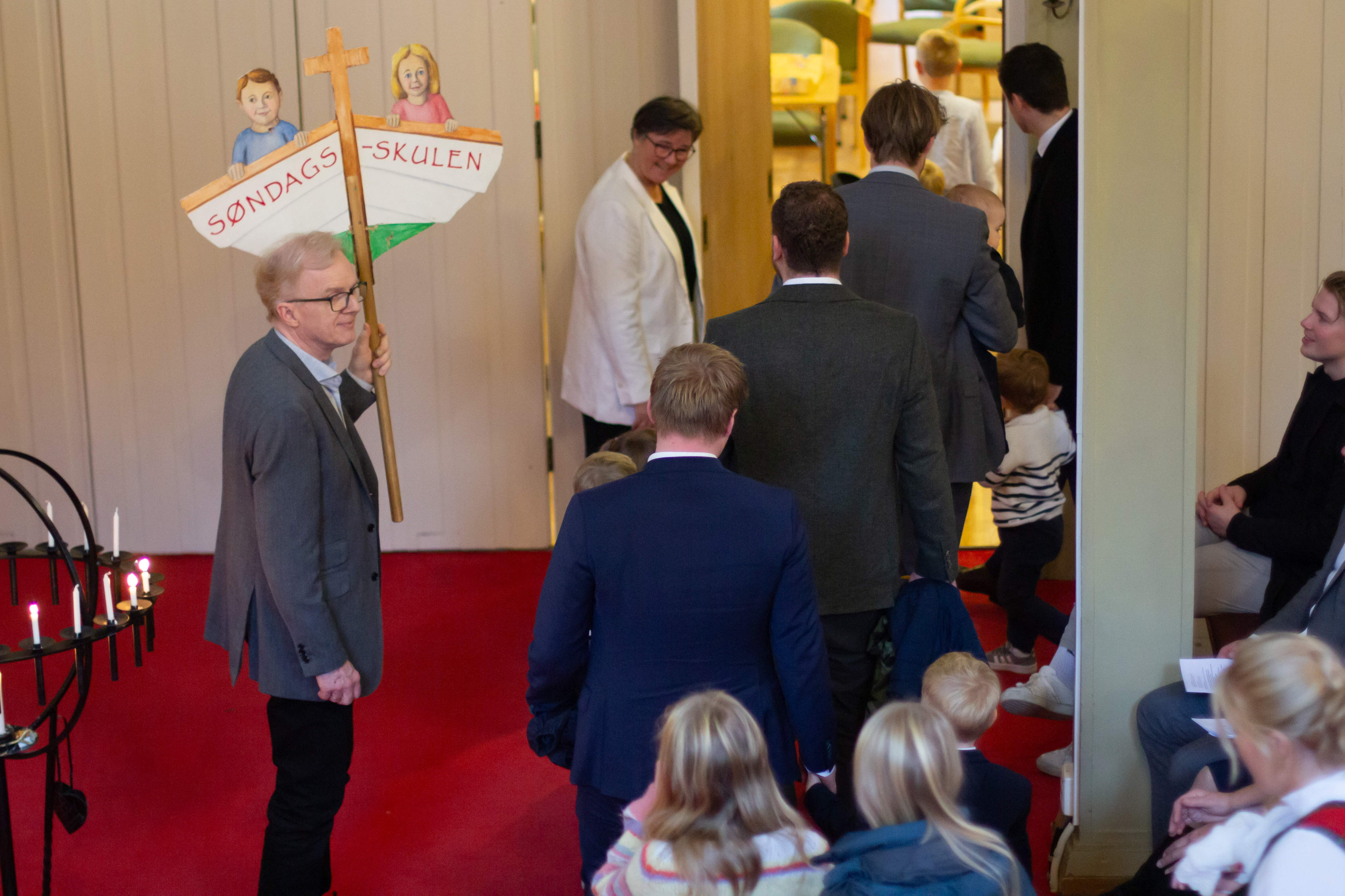 En mann holder et skilt, på skiltet er det en båt med to barn og "søndagsskule" skrevet under de. Barn og voksne er på vei ut av kirkerommet. 