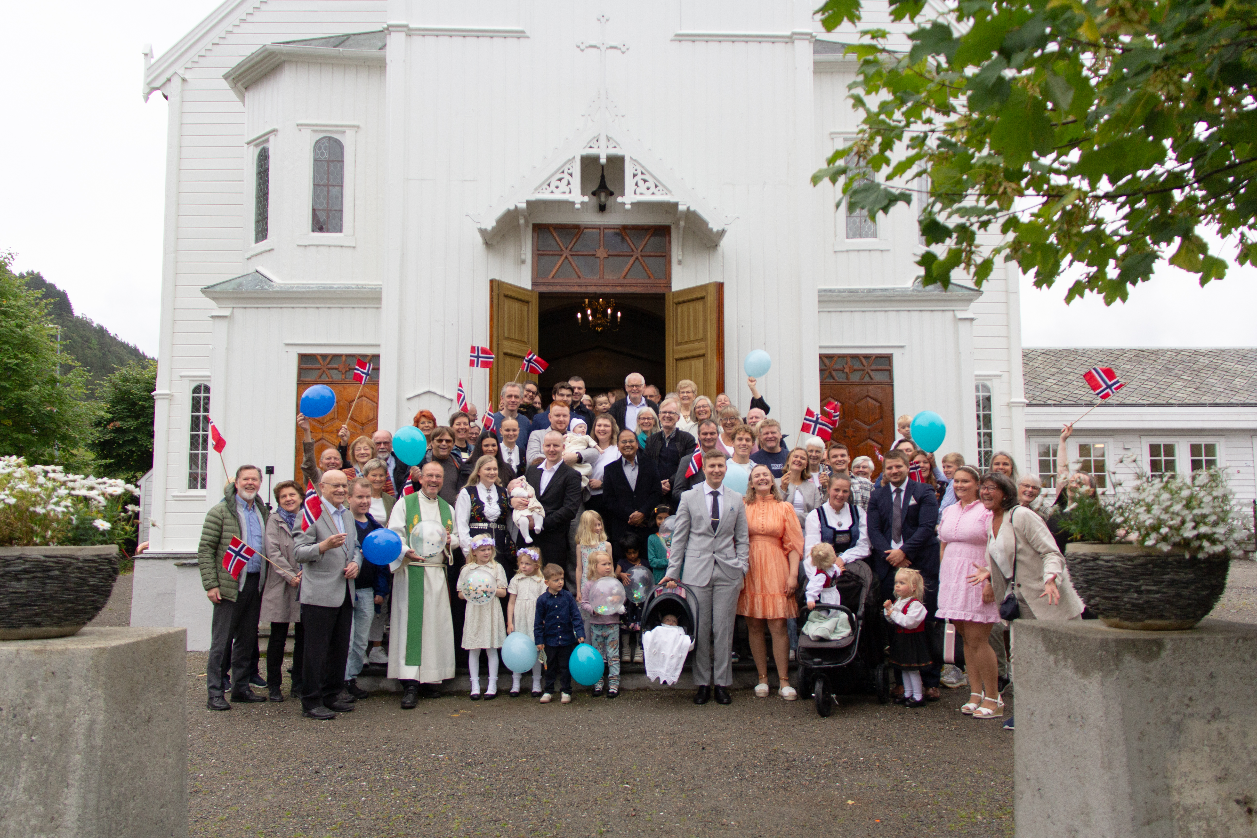 Mange mennesker i alle aldre står i kirketrappen, noen er dåpsfamilier. Noen holder ballonger og andre flagg, de jubler og er glad