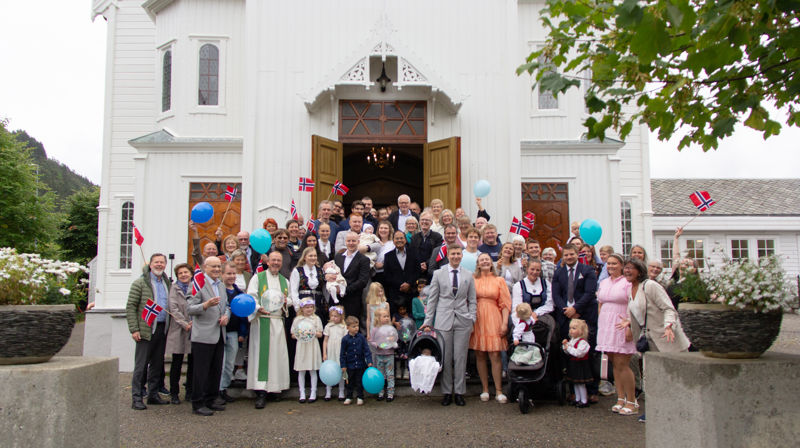 Mange mennesker i alle aldre står i kirketrappen, noen er dåpsfamilier. Noen holder ballonger og andre flagg, de jubler og er glad