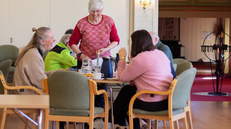 En dame står ved et bord og smiler og ler sammen med andre som sitter ved bordet