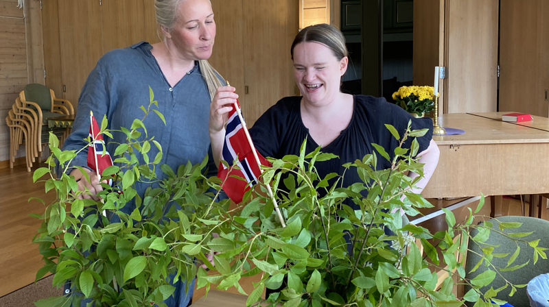 To damer stå og lager til vaser på et bord, oppi vasene er grønne grener og norske flagg