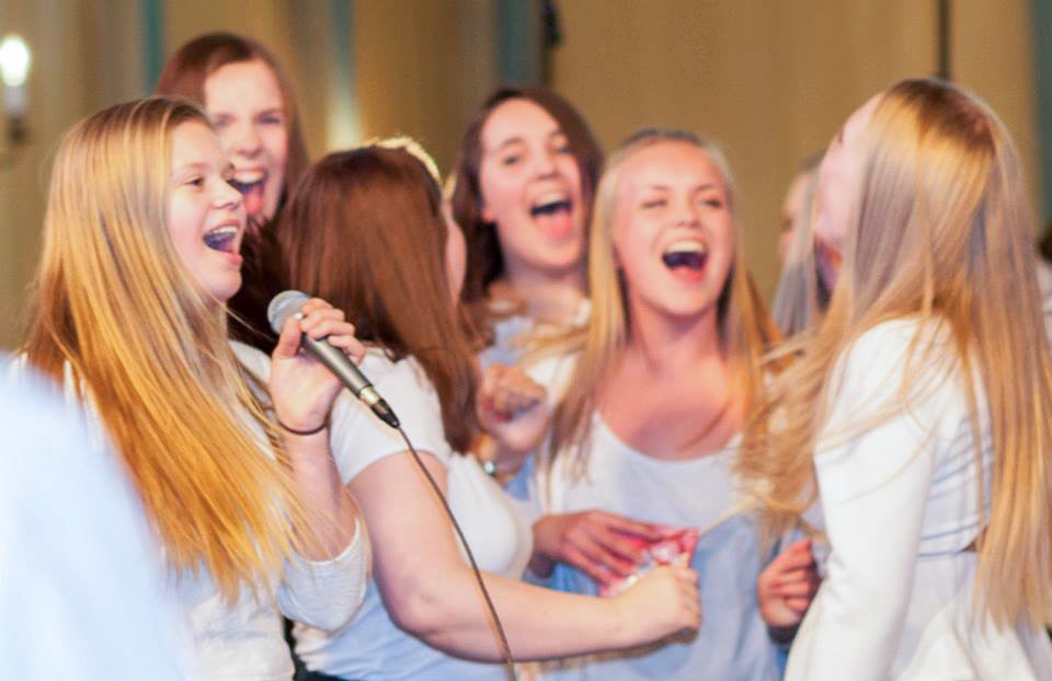 En tenåringsjente synger i en mikrofon og flere jenter står i bakgrunnen og synger smilende 