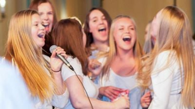 En tenåringsjente synger i en mikrofon og flere jenter står i bakgrunnen og synger smilende 