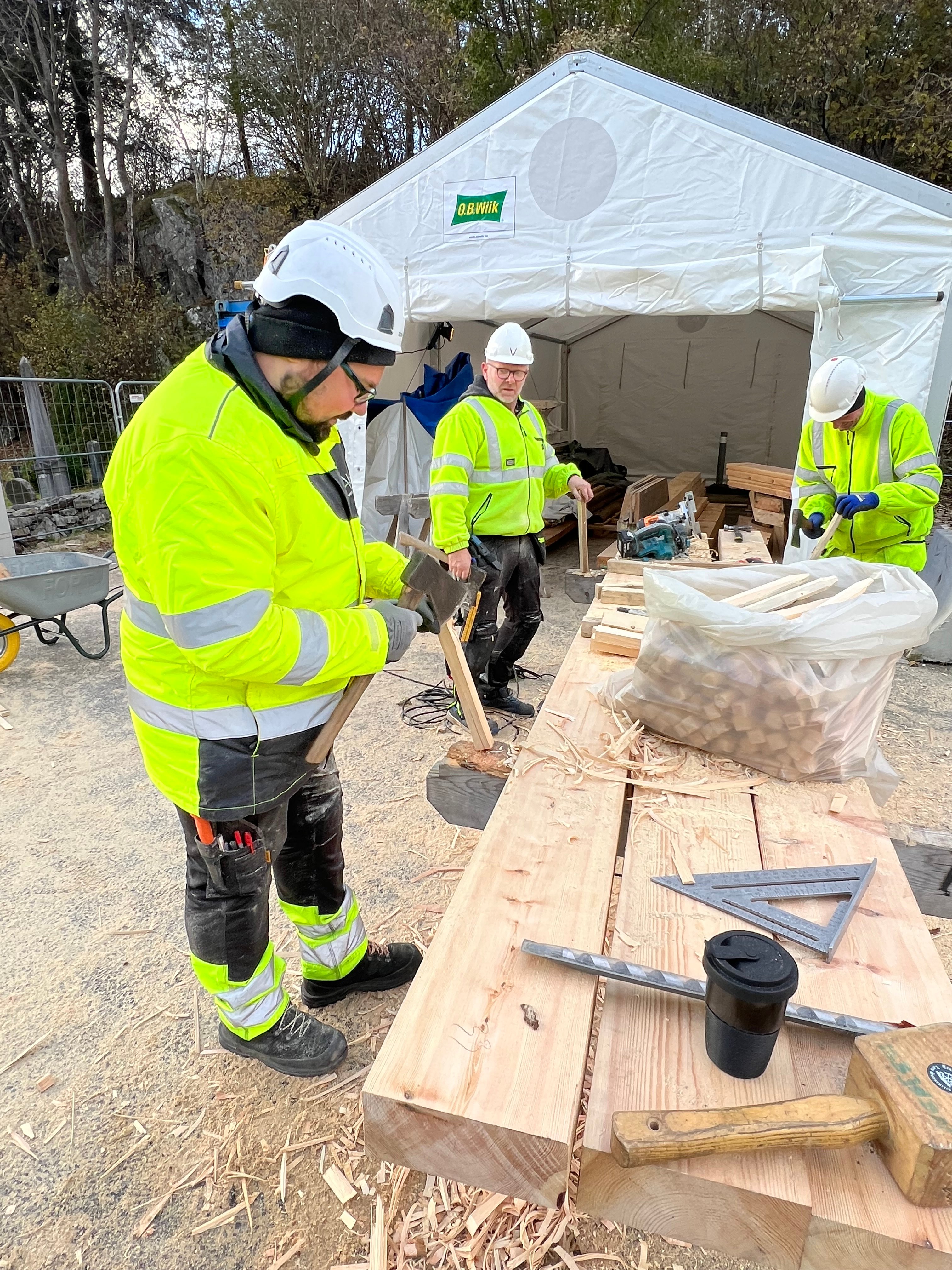 Tre arbeidarar står ute rundt eit arbeidsbord og lagar tre-pinnar ved hjelp av øks