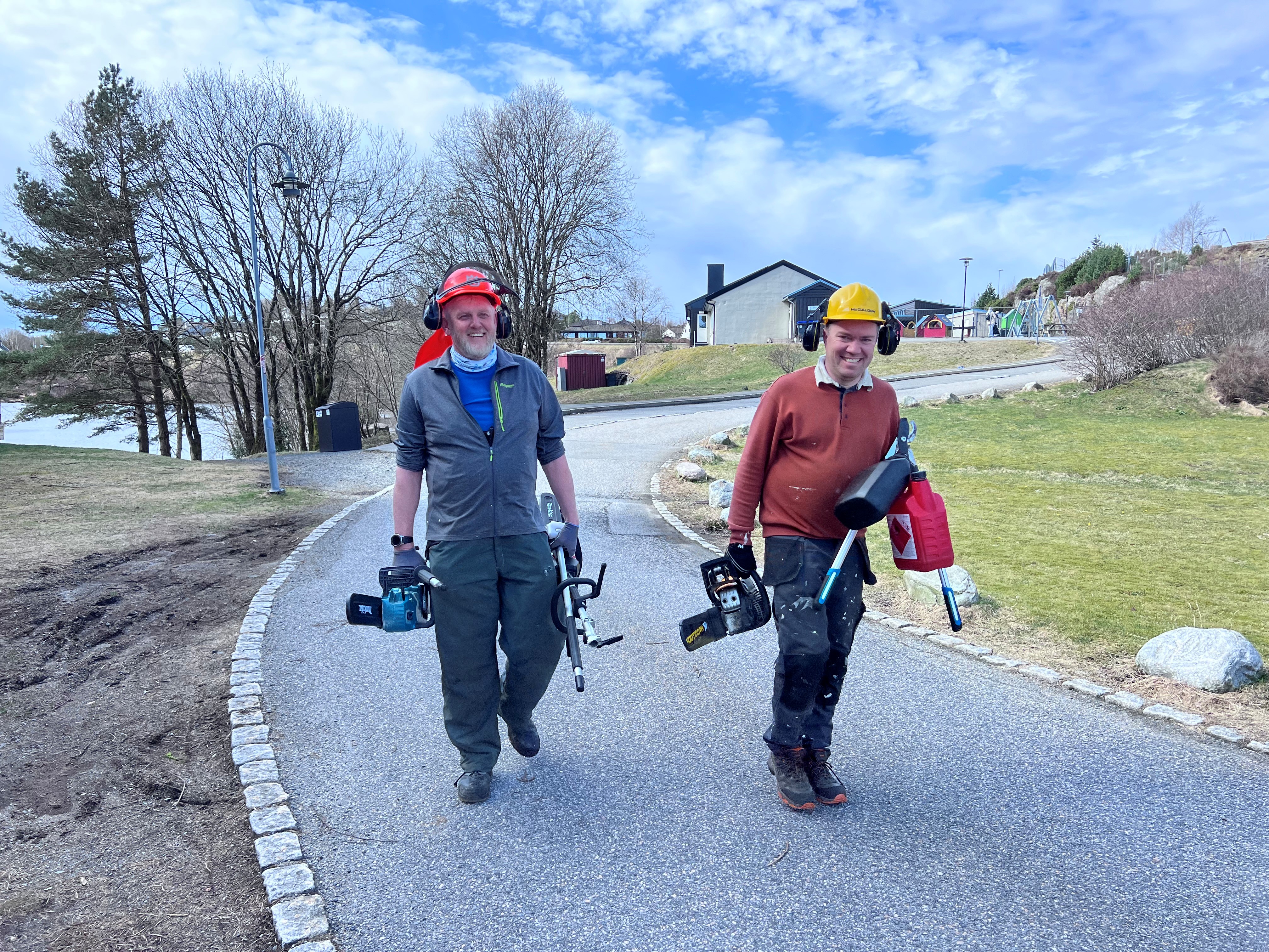 To menn kommer gående smilende, de har hjelm på hodet og verktøy i hendene