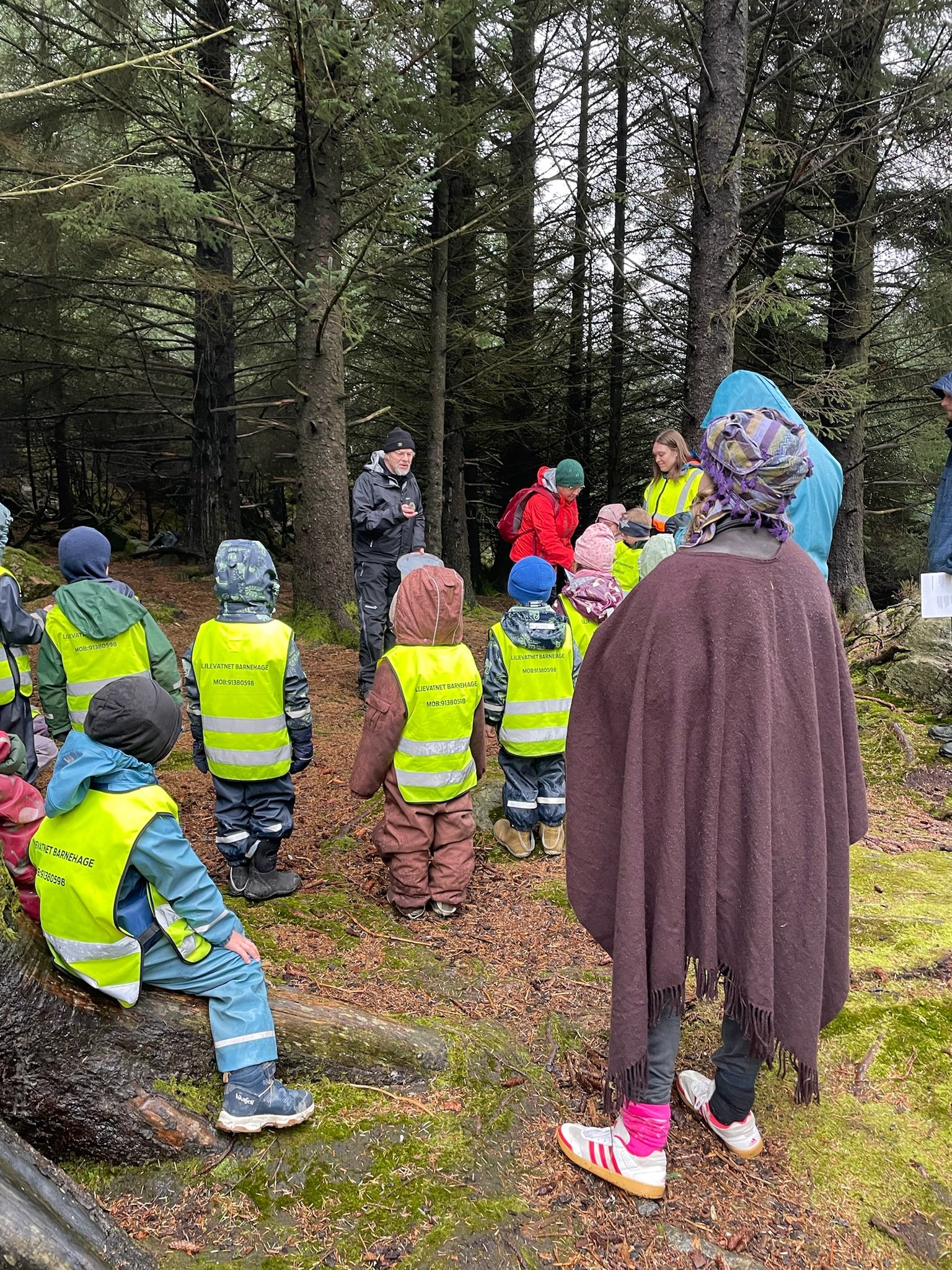 Barnehagebarn i refleksvester og vaksne står inne i en åpning i en skog, en mann holder et ark og forteller