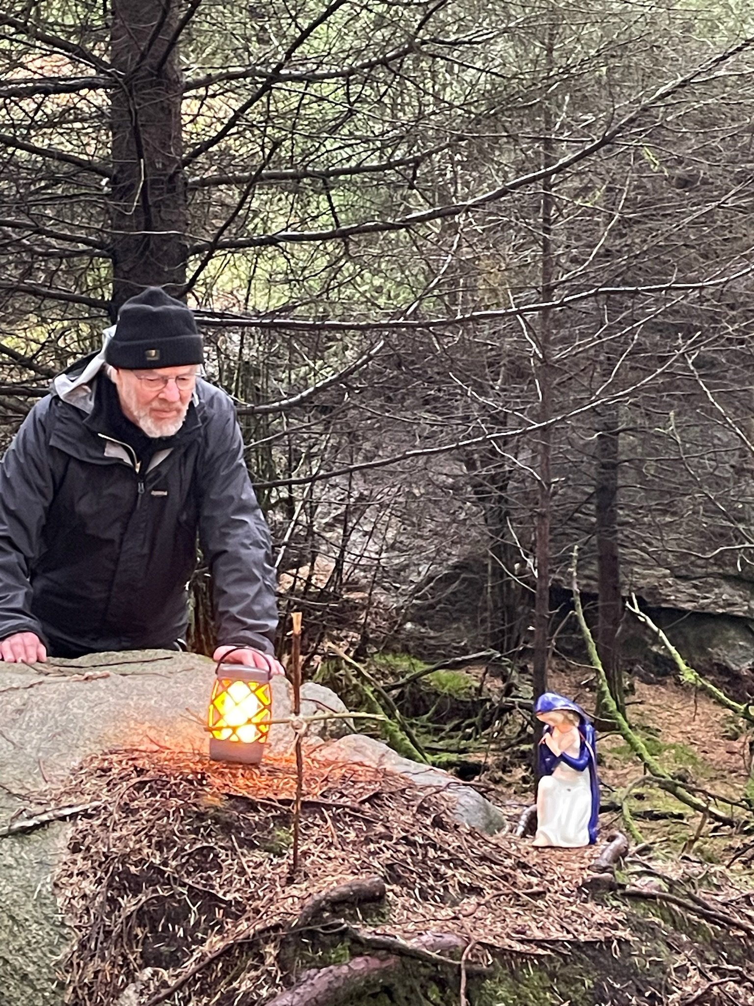 En mann står bak en stor stein, oppå steinen står en lykt som lyser, et kors laget av pinner og en figur av Maria