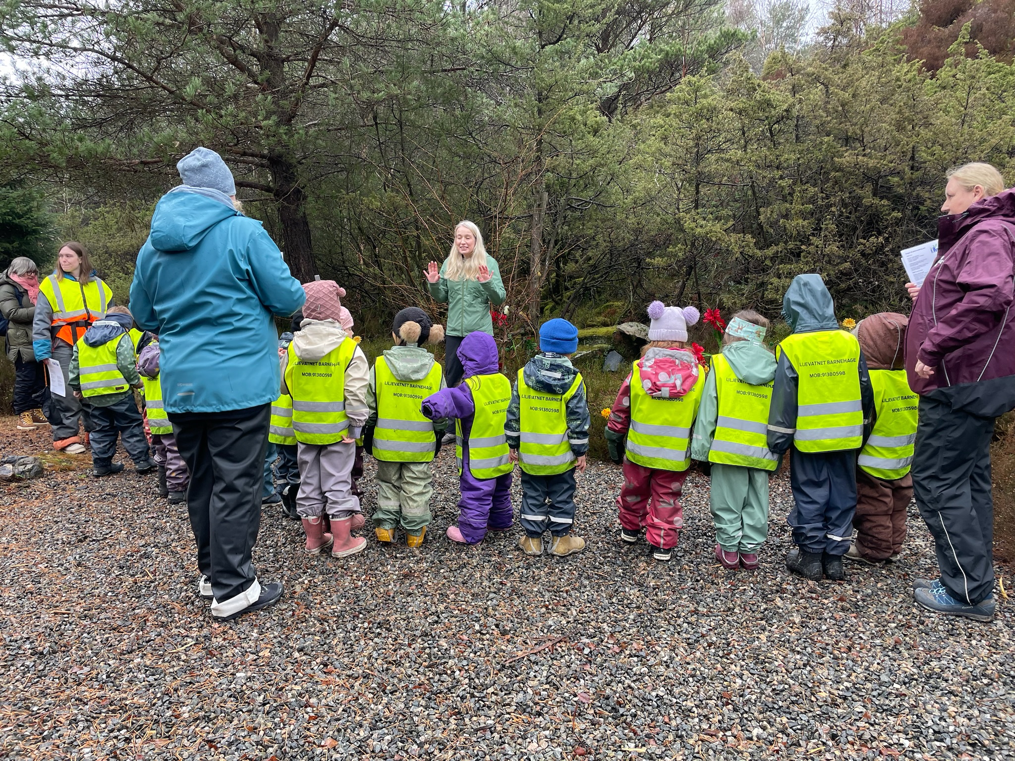 Ute på en grusvei står barnehagebarn med refleksvester i en halvsirkel, framme står en dame og forteller noe