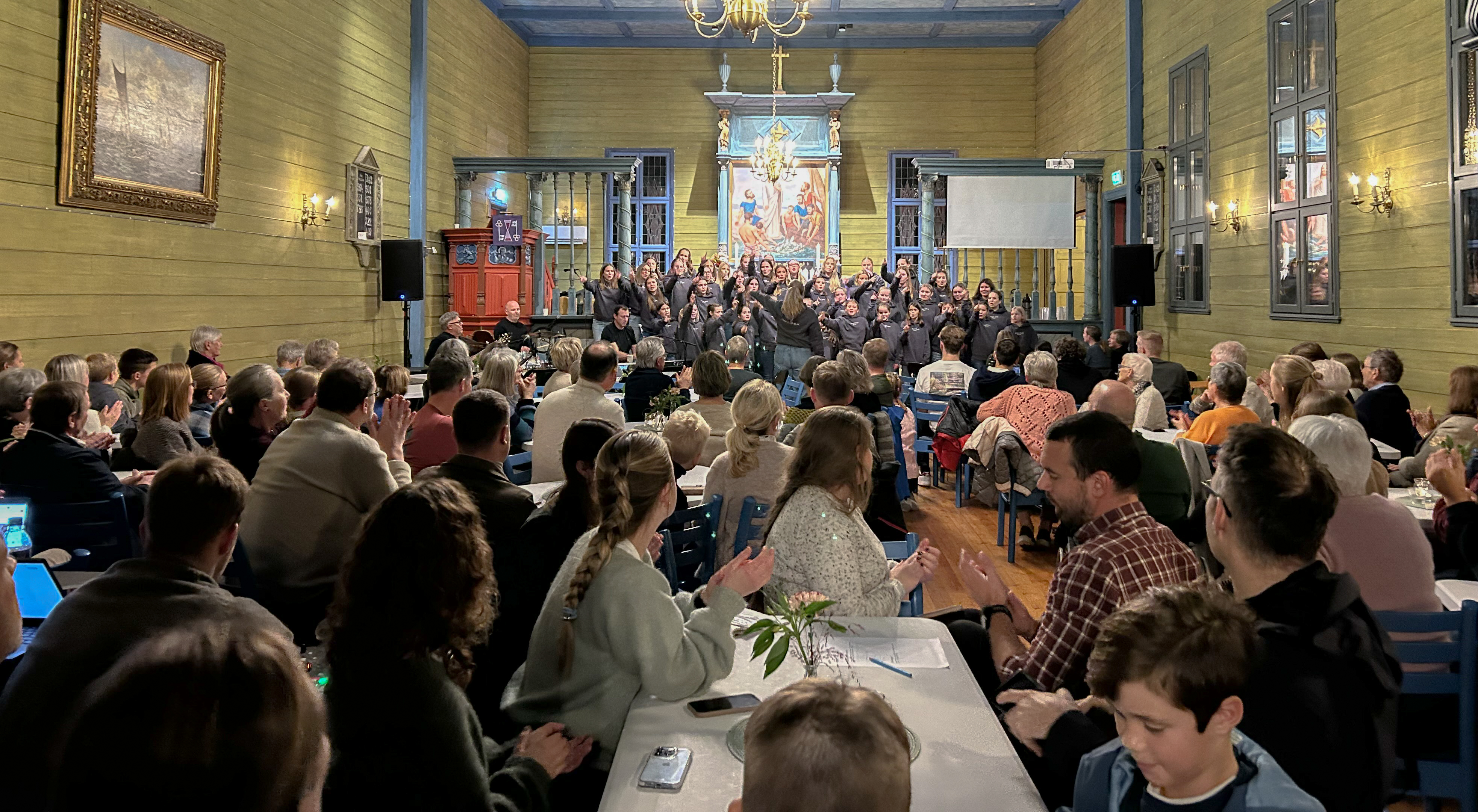 Mennesker sitter i kirken rundt bord, fremme står et stort kor med tenåringsjenter i svarte genser og synger