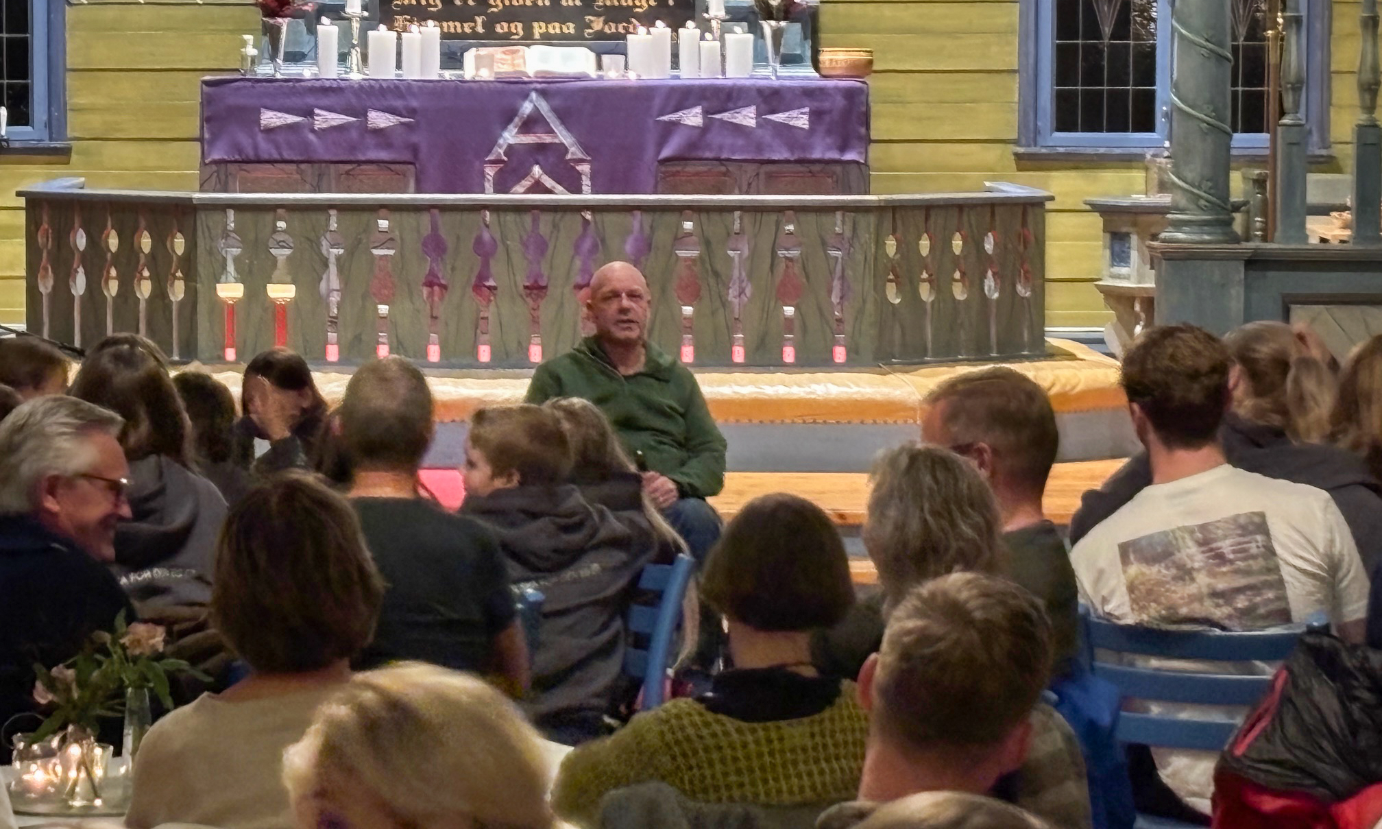 En mann i grønn genser sitter fremme i kirken og forteller til publikum