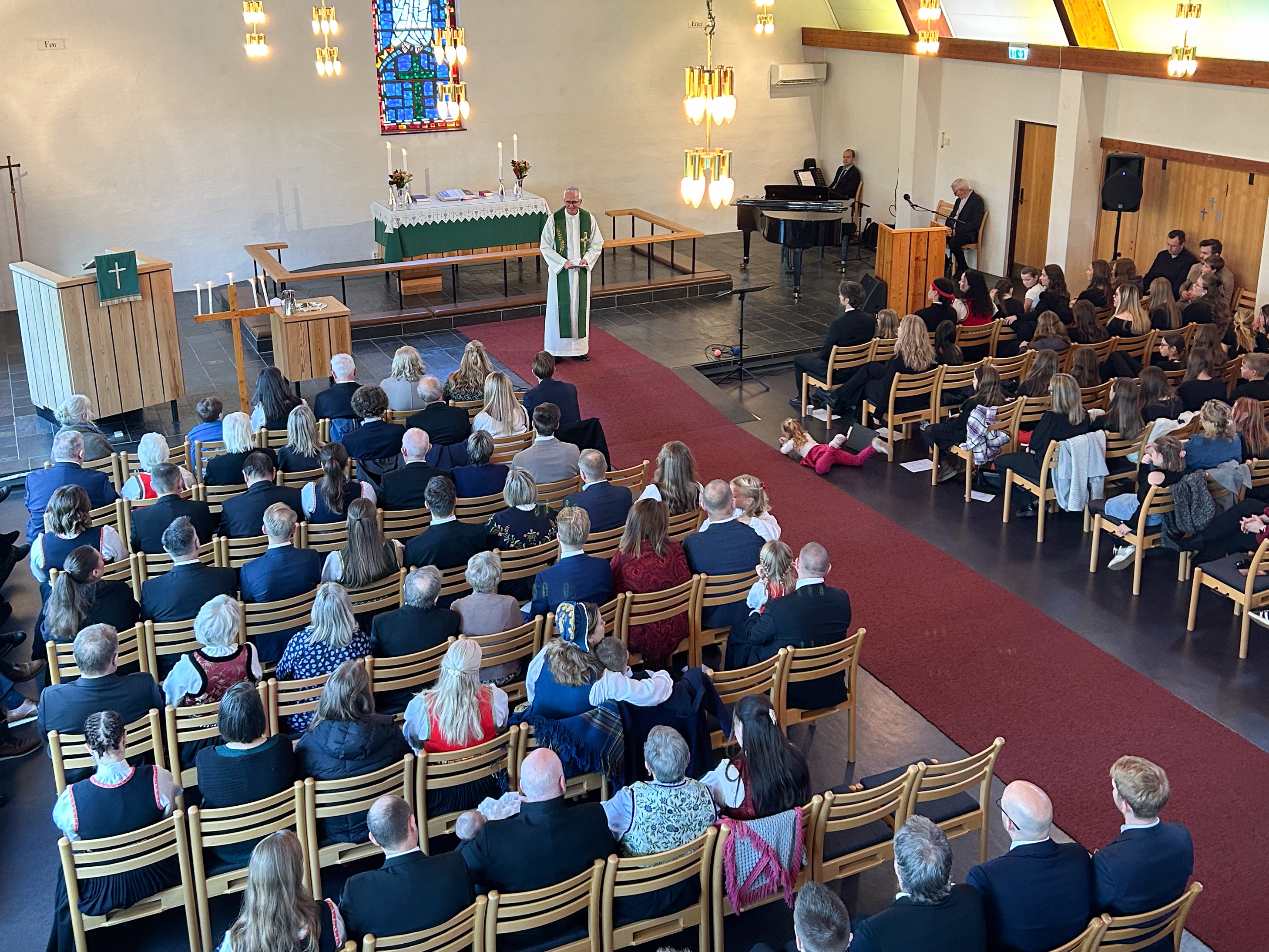 inne i en kirke står presten fremme og taler, mennesker sitter tett i tett på stoler