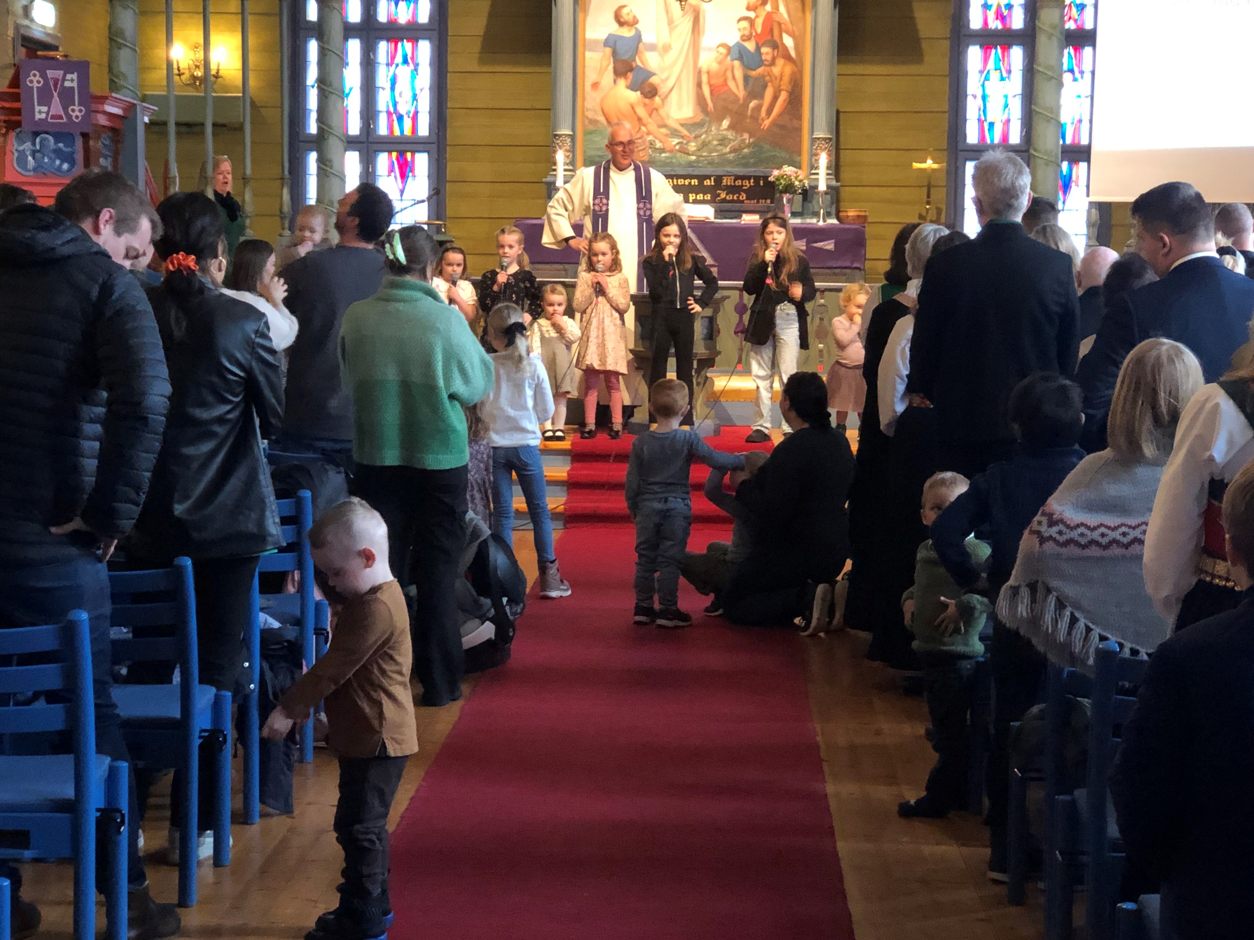 Gudsteneste i Blomvåg kyrkje. Kyrkjelyden står og syng og framme står presten saman med barn som syng i mikrofon. På det raude teppet i midtgangen sitt fleire barn. 
