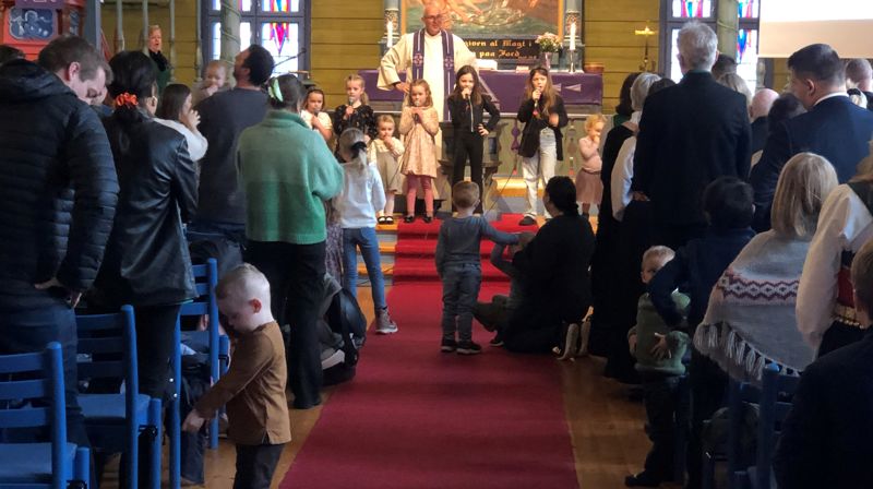 Gudsteneste i Blomvåg kyrkje. Kyrkjelyden står og syng og framme står presten saman med barn som syng i mikrofon. På det raude teppet i midtgangen sitt fleire barn.