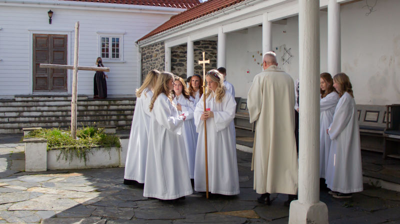 Konfirmanter i hvite kapper står ute i borggården med presten, snart klar til å gå inn i prosesjon. En jente holder prosesjonskorset. Det er fint vær.