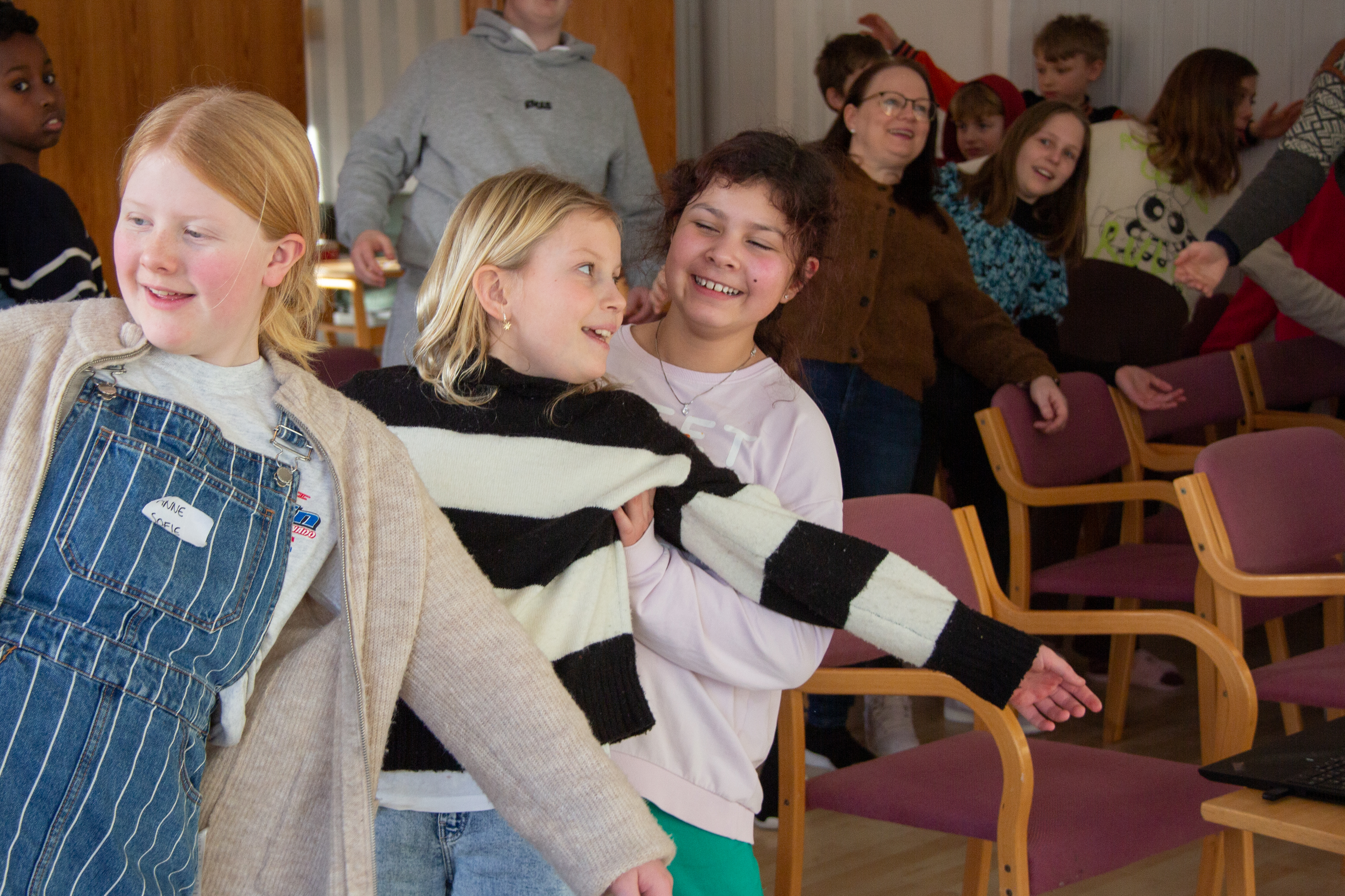 Tre jenter står og lener seg bak mot hverandre og smiler. Man kan se andre barn, voksne og ungdommer i stolrader bak de i samme bevegelse.