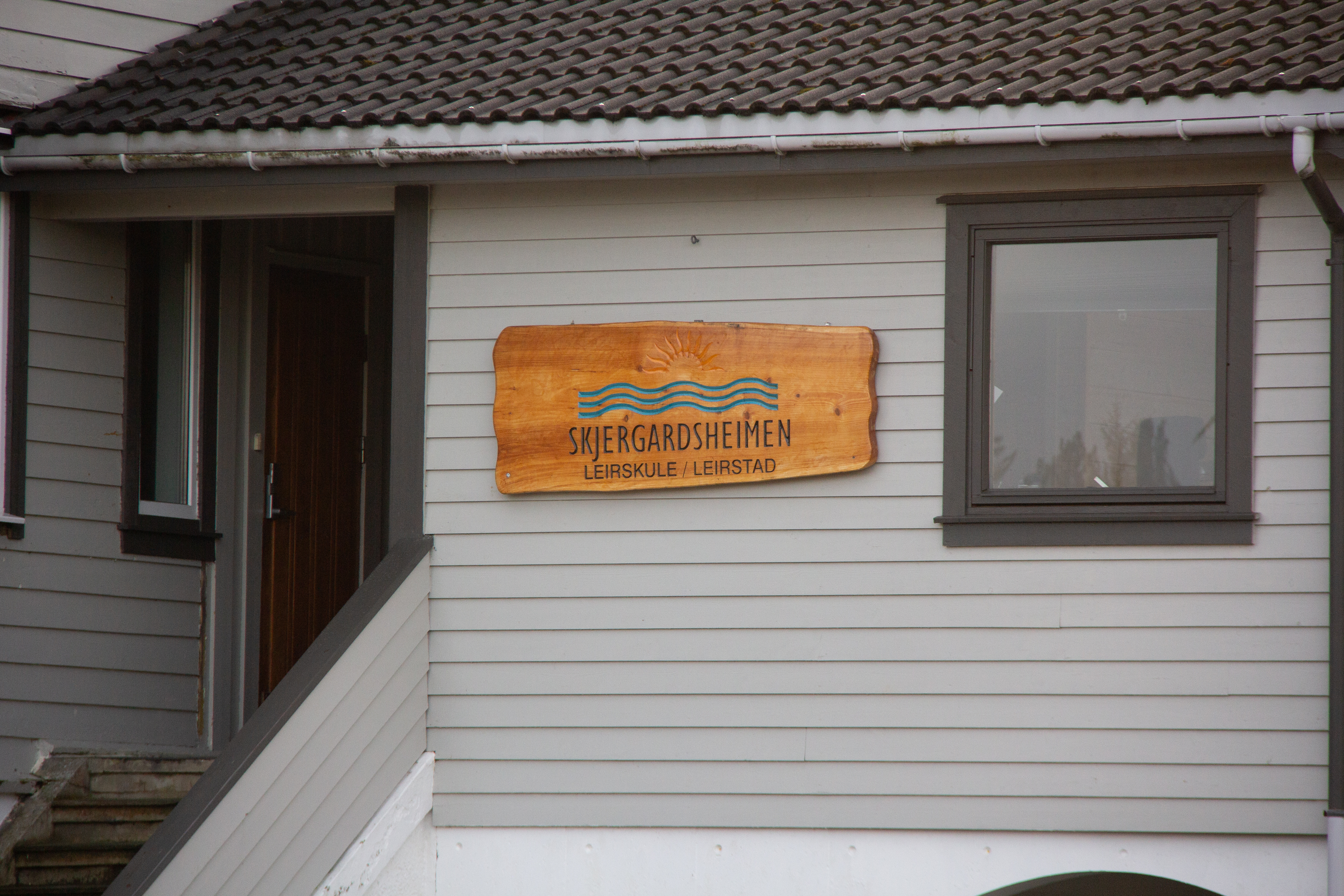 Bilde av en hvit yttervegg av et hus, på veggen henger det et skilt av tre med Skjergardsheimen sin logo skjert inn.