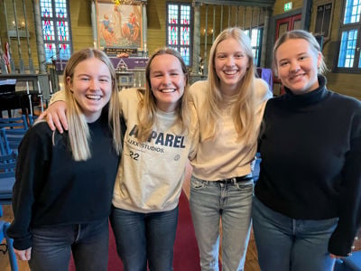 Fire ungdommer, jenter, står samlet og smiler til kameraet. De er inne i Blomvåg kyrkje. 