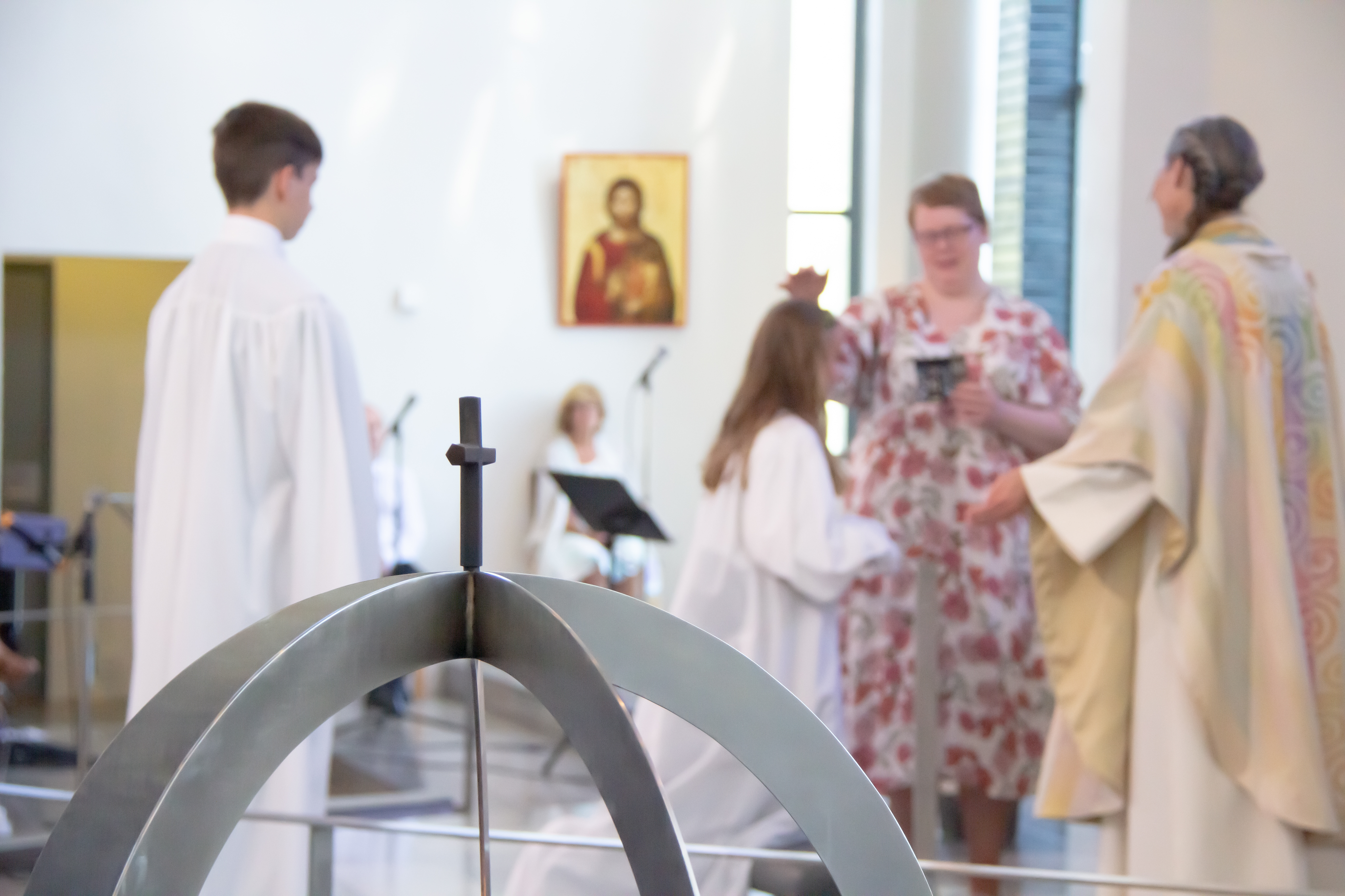 Fokus på kors over lysglobe, uklart i bakgrunnen blir en konfirmant bedt for av konfirmantlærar og prest