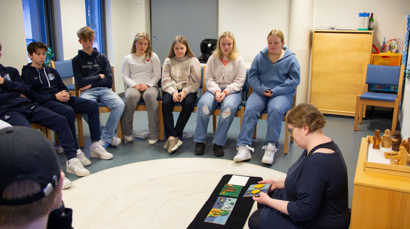 Ungdommer sitter i en halvsirkel og ser på en konfirmantlærer som viser frem en historie