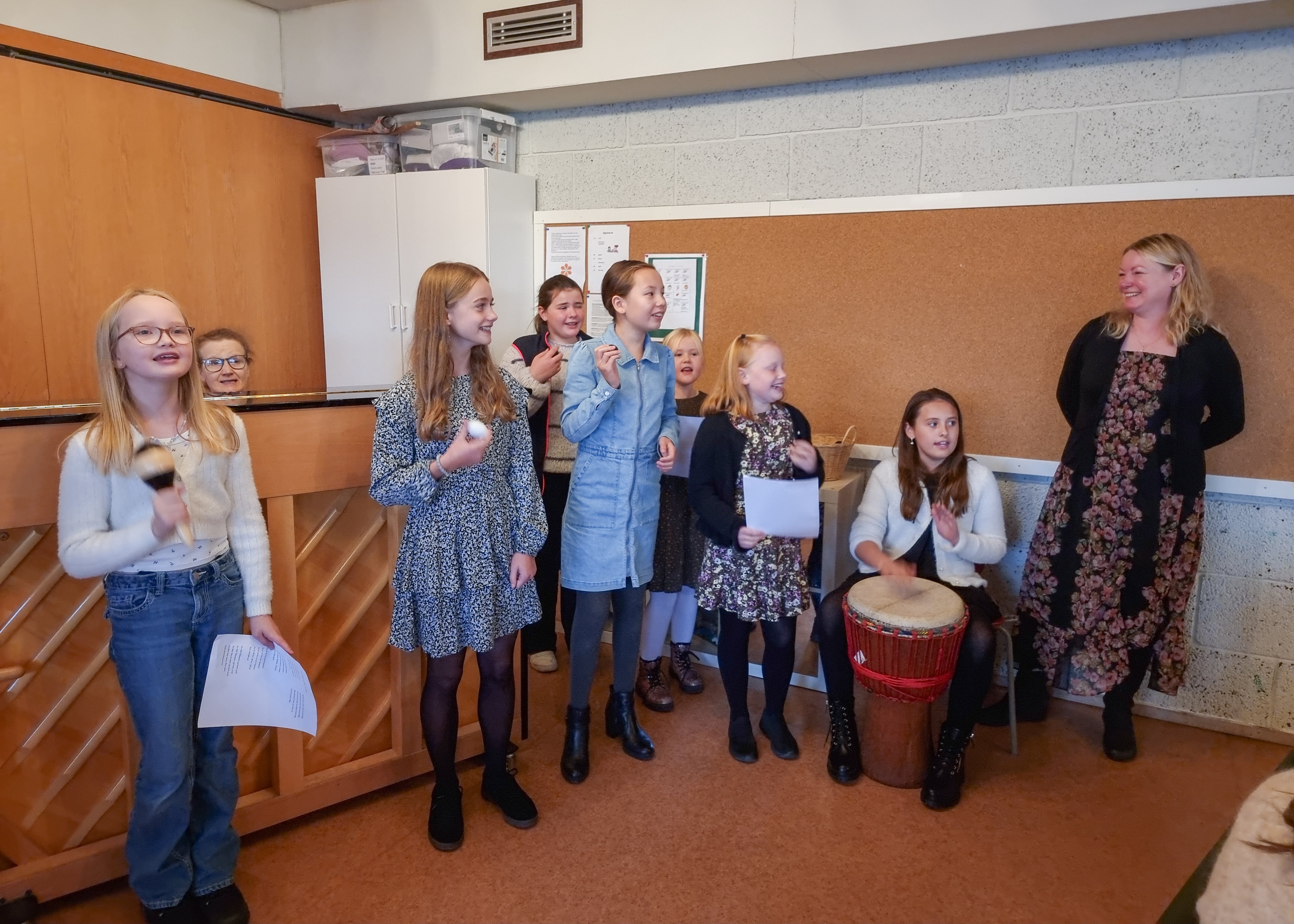 tweensjenter synger og spiller trommeinstrument, til høyre står en dame og er rørt