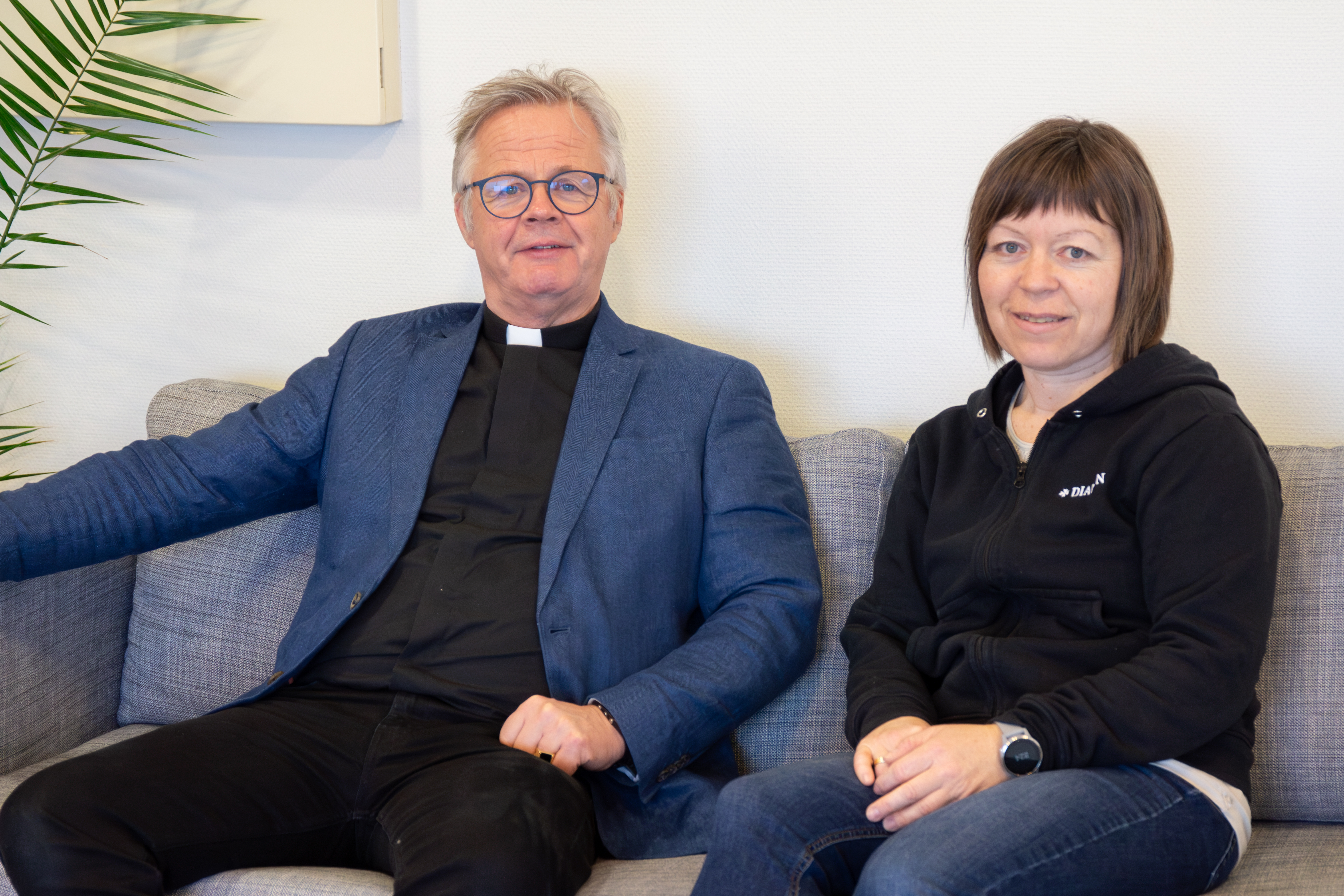 En prest og en diakon sitter på en sofa og ser inn i kameraet