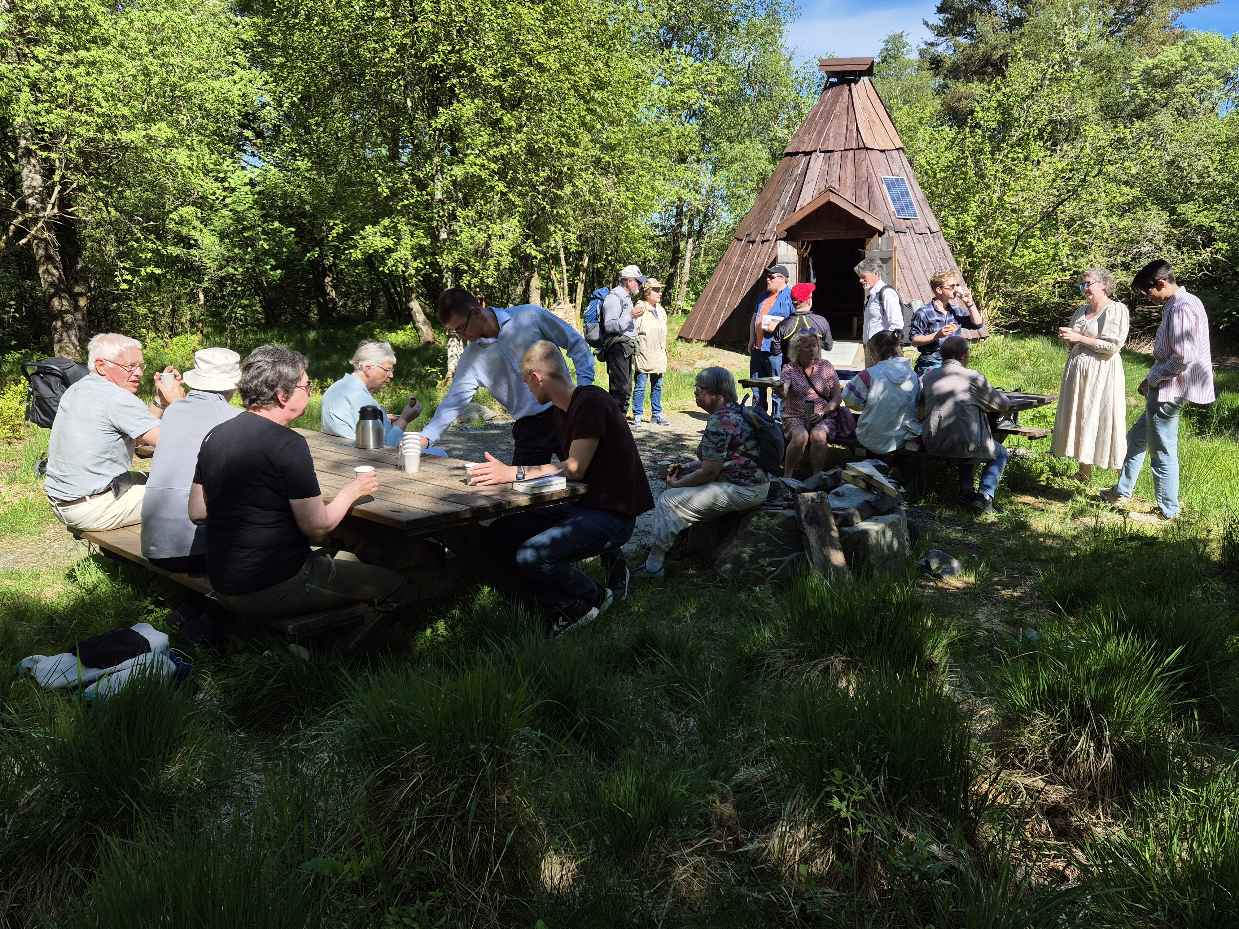 Ute i naturen sitter og står 18 mennesker rundt piknikkbord, i bakgrunnen står en lavvo laget av tre.  Det er grønt og sommerlig i naturen og solen skinner