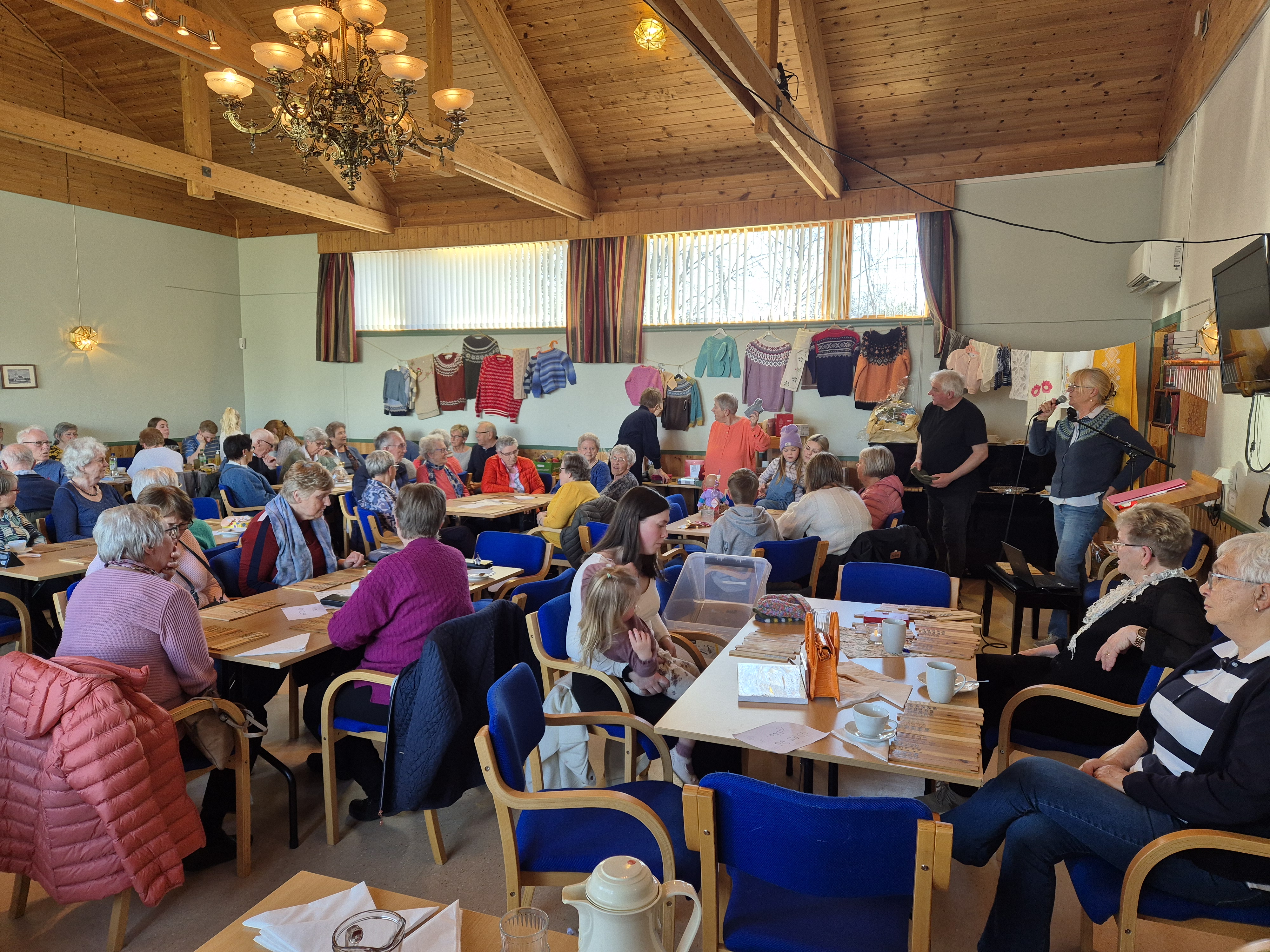 Inne i kyrkjelydshuset er det fullt av mennesker rundt bord, på veggen hhenger mstrikkede gensere, det er basar