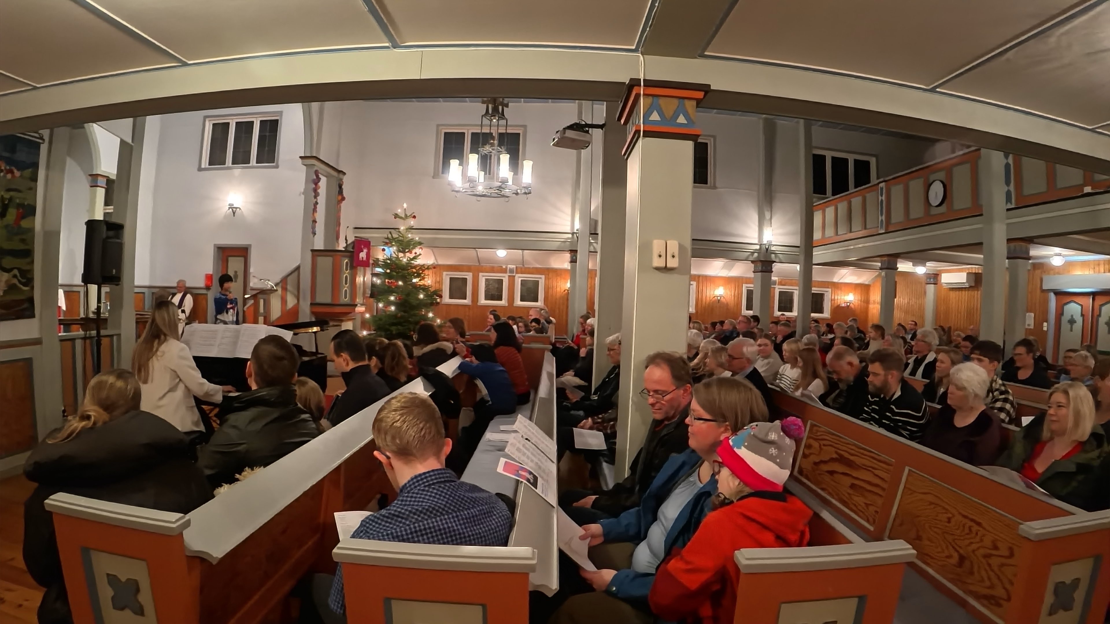 Kyrkjegjengarar sit i benkradene inni kyrkja, til venstre kan ein sjå eit juletre
