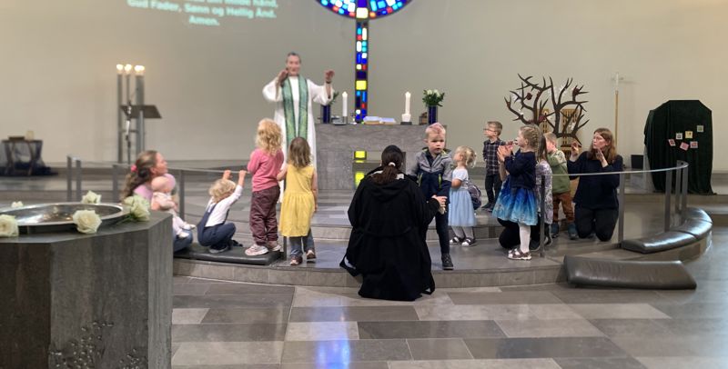 Presten står framme i kyrkja saman med mange 4-åringar og nokre foreldre, ho lagar bevegelser til ein song